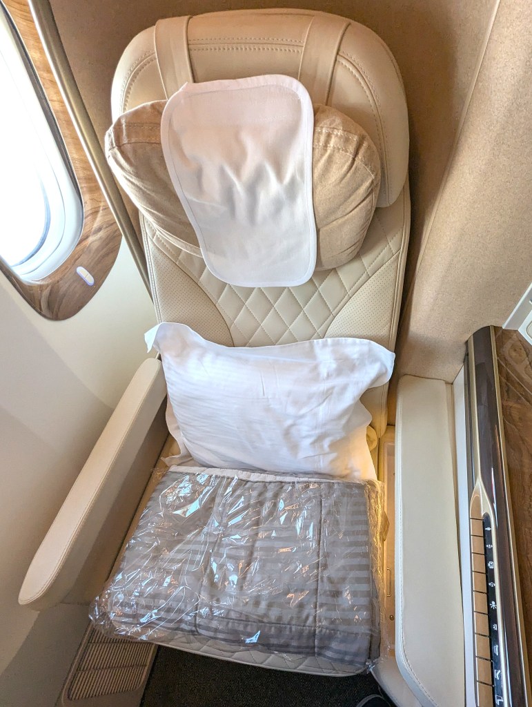 Business class seat on Emirates Airlines featuring a comfortable headrest, pillow, and a blanket wrapped in plastic, positioned next to a window.