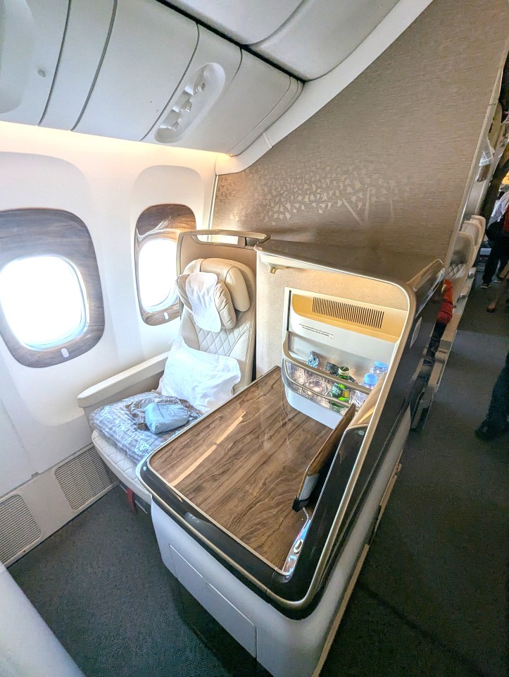 Interior view of an Emirates Airlines business class seat with a large window, comfortable seat, and a wooden table with amenities.