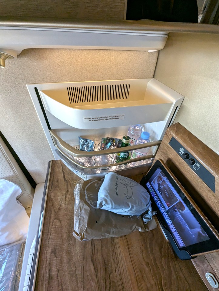 Interior view of an Emirates Airlines business class seat showing mini bar or storage compartment with water bottles, snacks, and an entertainment screen on a wooden table.
