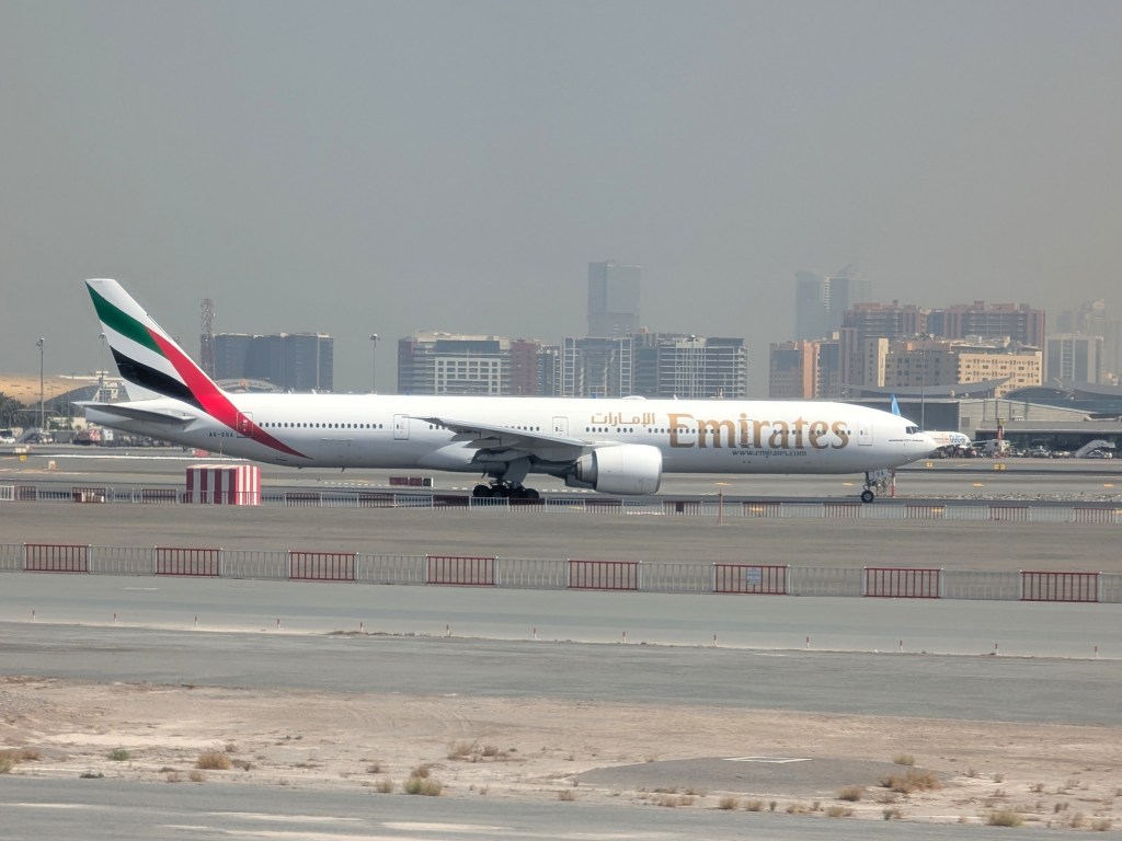 An Emirates Airlines Boeing 777-300 aircraft on a runway, with city buildings in the background.